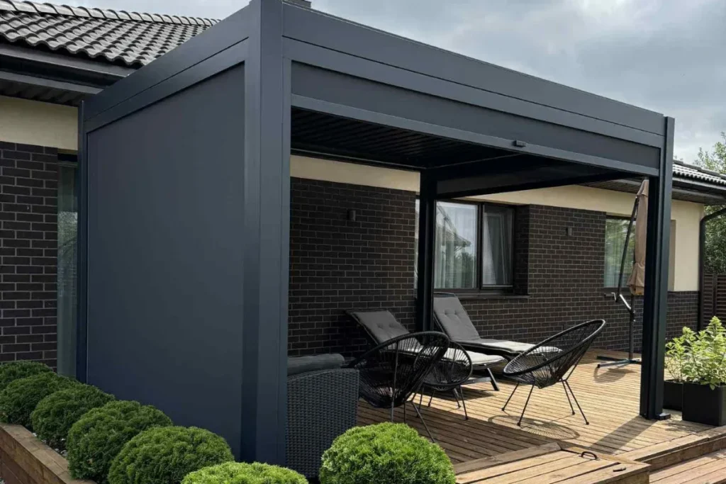 Medinė terasa su modernia tamsaus aliuminio pergola ir poilsio baldais prie namo.