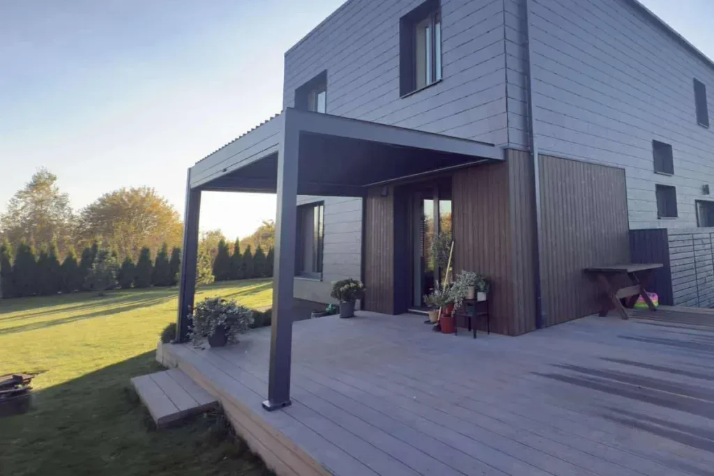 Moderni tamsi pergola pritvirtinta prie dviejų aukštų namo, su plačiu mediniu kiemo deniu ir dekoratyviniais augalais.