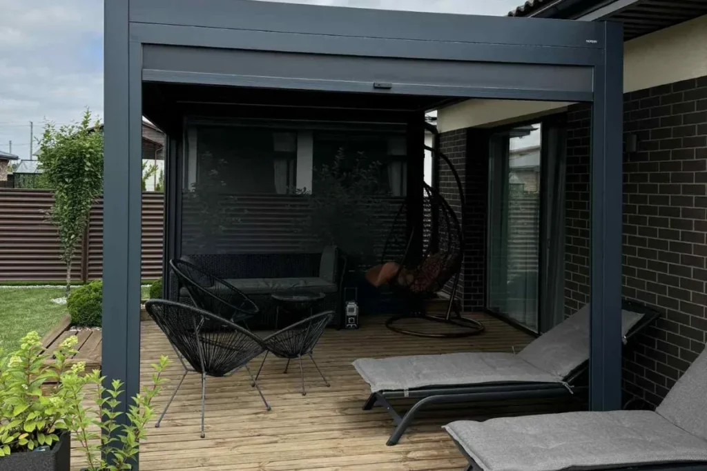Moderni pergola su uždanga virš terasos, kurioje įrengta lauko poilsio zona su gultais ir kėdėmis.