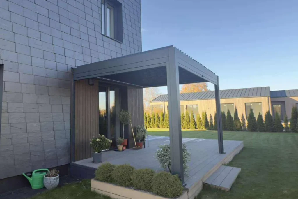 Tamsi pergola iš šono, sumontuota prie šiuolaikiško fasado, su terasa ir tvarkingai sutvarkyta veja.