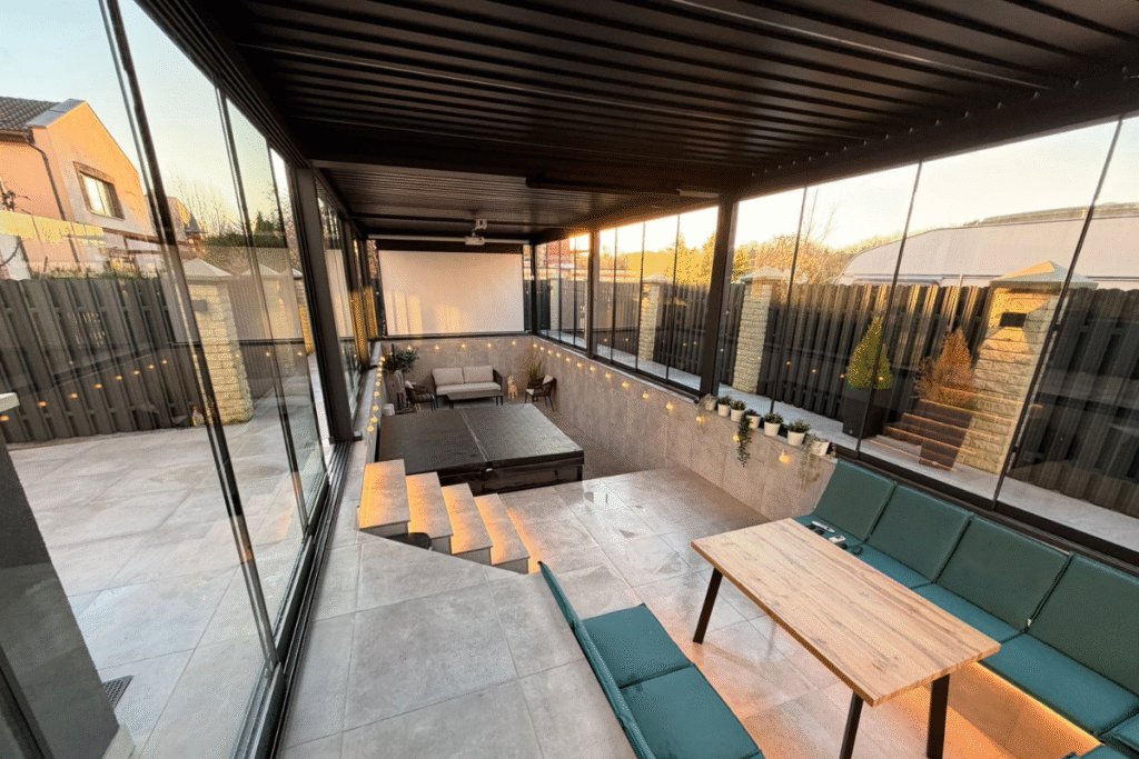 Stiklinėmis sienelėmis uždara pergola su tamsiu stogu ir integruotu apšvietimu, įrengta su lauko baldais ir apdaila.