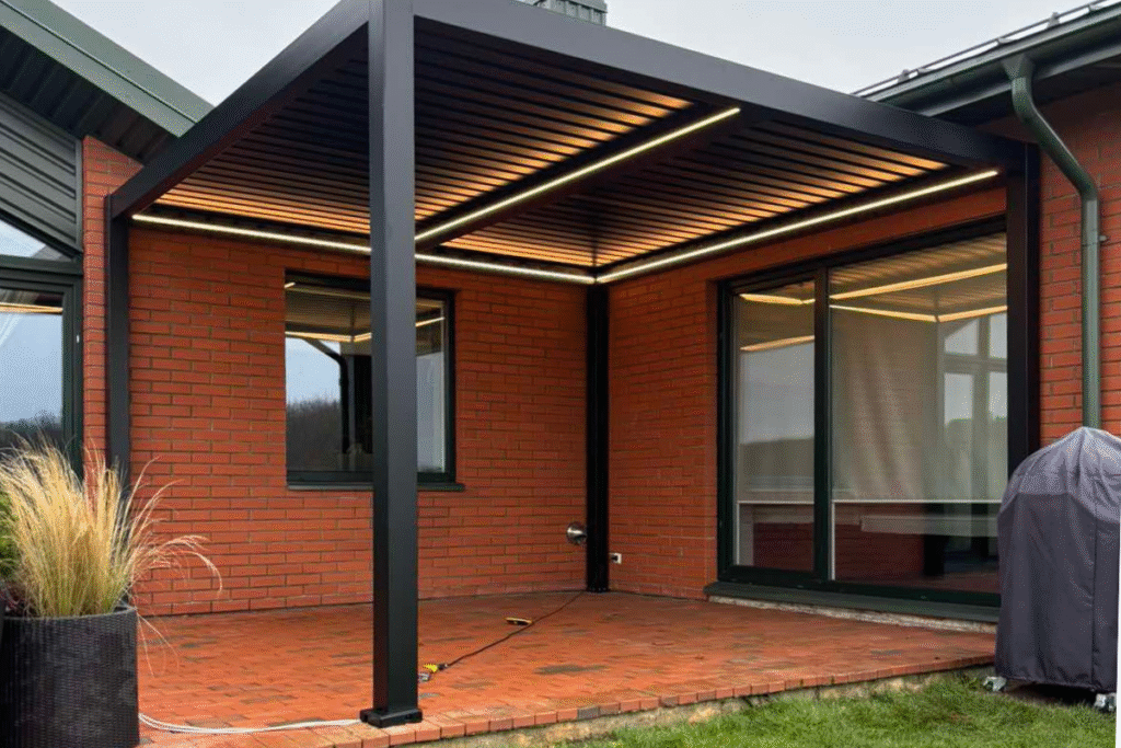 C4S 4x4 pergola su apšvietimu, pritvirtinta prie raudonų plytų namo, su atviru stogu ir terasa.